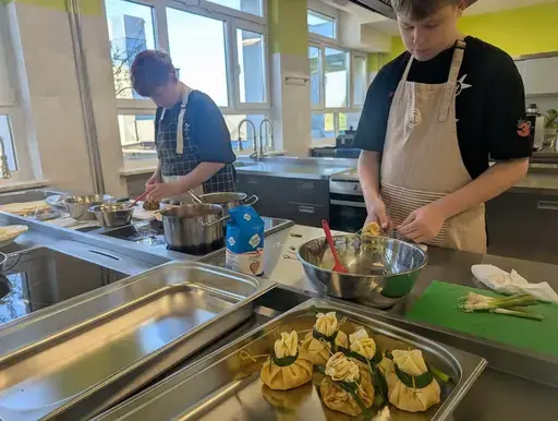 
                                                    Uczniowie Zespołu Szkół Zawodowych nr 2 w Dęblinie w fartuchach smażą i nakładają naleśniki w szkolnej pracowni gastronomicznej oraz przy stoiskach nakrytych zielonymi obrusami, z czarnymi banerami z nazwami dań, udekorowanych różowymi i białymi balonami; na stołach porcje naleśników z dodatkami na białych talerzykach.
                                                