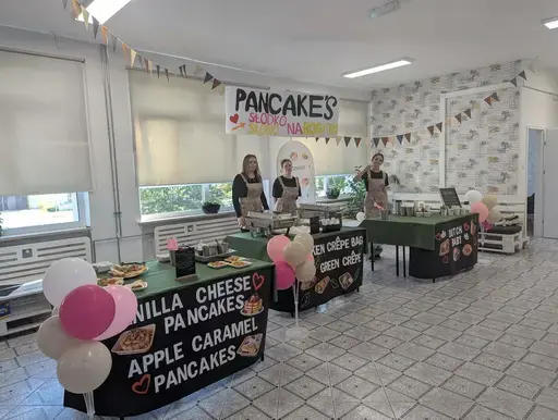 
                                                    Uczniowie Zespołu Szkół Zawodowych nr 2 w Dęblinie w fartuchach smażą i nakładają naleśniki w szkolnej pracowni gastronomicznej oraz przy stoiskach nakrytych zielonymi obrusami, z czarnymi banerami z nazwami dań, udekorowanych różowymi i białymi balonami; na stołach porcje naleśników z dodatkami na białych talerzykach.
                                                