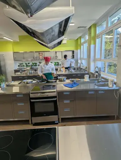 
                                                    Uczniowie Zespołu Szkół Zawodowych nr 2 w Dęblinie w fartuchach smażą i nakładają naleśniki w szkolnej pracowni gastronomicznej oraz przy stoiskach nakrytych zielonymi obrusami, z czarnymi banerami z nazwami dań, udekorowanych różowymi i białymi balonami; na stołach porcje naleśników z dodatkami na białych talerzykach.
                                                