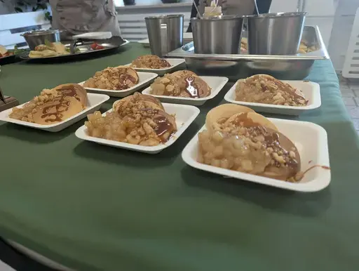 
                                                    Uczniowie Zespołu Szkół Zawodowych nr 2 w Dęblinie w fartuchach smażą i nakładają naleśniki w szkolnej pracowni gastronomicznej oraz przy stoiskach nakrytych zielonymi obrusami, z czarnymi banerami z nazwami dań, udekorowanych różowymi i białymi balonami; na stołach porcje naleśników z dodatkami na białych talerzykach.
                                                