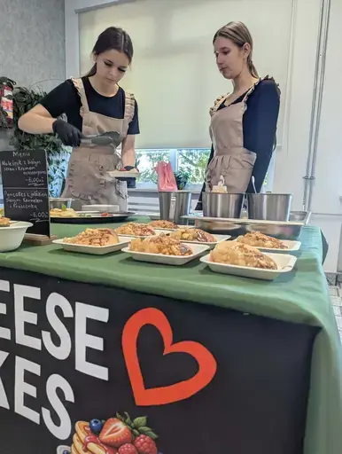 
                                                    Uczniowie Zespołu Szkół Zawodowych nr 2 w Dęblinie w fartuchach smażą i nakładają naleśniki w szkolnej pracowni gastronomicznej oraz przy stoiskach nakrytych zielonymi obrusami, z czarnymi banerami z nazwami dań, udekorowanych różowymi i białymi balonami; na stołach porcje naleśników z dodatkami na białych talerzykach.
                                                