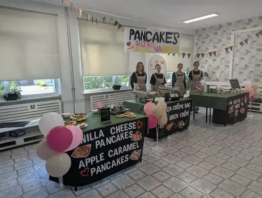 
                                                    Uczniowie Zespołu Szkół Zawodowych nr 2 w Dęblinie w fartuchach smażą i nakładają naleśniki w szkolnej pracowni gastronomicznej oraz przy stoiskach nakrytych zielonymi obrusami, z czarnymi banerami z nazwami dań, udekorowanych różowymi i białymi balonami; na stołach porcje naleśników z dodatkami na białych talerzykach.
                                                