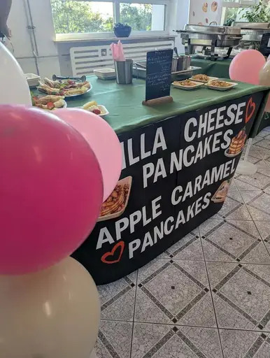 
                                                    Uczniowie Zespołu Szkół Zawodowych nr 2 w Dęblinie w fartuchach smażą i nakładają naleśniki w szkolnej pracowni gastronomicznej oraz przy stoiskach nakrytych zielonymi obrusami, z czarnymi banerami z nazwami dań, udekorowanych różowymi i białymi balonami; na stołach porcje naleśników z dodatkami na białych talerzykach.
                                                