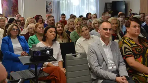 
                                                    Uczestnicy powiatowych obchodów Światowego Dnia Zdrowia Psychicznego w sali Ośrodka Wsparcia w Leopoldowie: goście i publiczność na krzesłach, prelegenci z mikrofonem przy ekranie projekcyjnym oraz występujący na tle granatowego banera z nazwą wydarzenia, ozdobionego kolorowymi jesiennymi liśćmi.
                                                