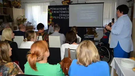 
                                                    Uczestnicy powiatowych obchodów Światowego Dnia Zdrowia Psychicznego w sali Ośrodka Wsparcia w Leopoldowie: goście i publiczność na krzesłach, prelegenci z mikrofonem przy ekranie projekcyjnym oraz występujący na tle granatowego banera z nazwą wydarzenia, ozdobionego kolorowymi jesiennymi liśćmi.
                                                