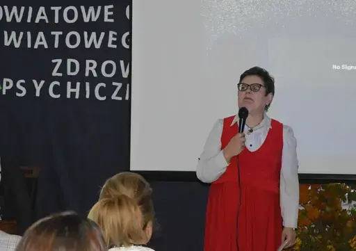 
                                                    Uczestnicy powiatowych obchodów Światowego Dnia Zdrowia Psychicznego w sali Ośrodka Wsparcia w Leopoldowie: goście i publiczność na krzesłach, prelegenci z mikrofonem przy ekranie projekcyjnym oraz występujący na tle granatowego banera z nazwą wydarzenia, ozdobionego kolorowymi jesiennymi liśćmi.
                                                