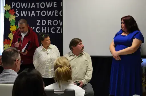
                                                    Uczestnicy powiatowych obchodów Światowego Dnia Zdrowia Psychicznego w sali Ośrodka Wsparcia w Leopoldowie: goście i publiczność na krzesłach, prelegenci z mikrofonem przy ekranie projekcyjnym oraz występujący na tle granatowego banera z nazwą wydarzenia, ozdobionego kolorowymi jesiennymi liśćmi.
                                                