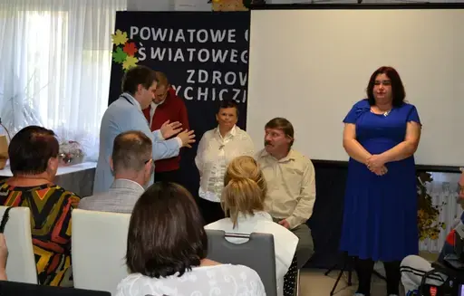 
                                                    Uczestnicy powiatowych obchodów Światowego Dnia Zdrowia Psychicznego w sali Ośrodka Wsparcia w Leopoldowie: goście i publiczność na krzesłach, prelegenci z mikrofonem przy ekranie projekcyjnym oraz występujący na tle granatowego banera z nazwą wydarzenia, ozdobionego kolorowymi jesiennymi liśćmi.
                                                