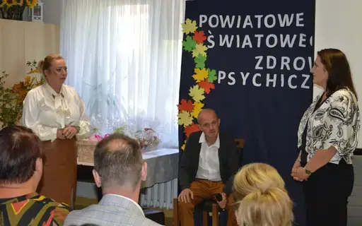 
                                                    Uczestnicy powiatowych obchodów Światowego Dnia Zdrowia Psychicznego w sali Ośrodka Wsparcia w Leopoldowie: goście i publiczność na krzesłach, prelegenci z mikrofonem przy ekranie projekcyjnym oraz występujący na tle granatowego banera z nazwą wydarzenia, ozdobionego kolorowymi jesiennymi liśćmi.
                                                