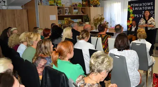 
                                                    Uczestnicy powiatowych obchodów Światowego Dnia Zdrowia Psychicznego w sali Ośrodka Wsparcia w Leopoldowie: goście i publiczność na krzesłach, prelegenci z mikrofonem przy ekranie projekcyjnym oraz występujący na tle granatowego banera z nazwą wydarzenia, ozdobionego kolorowymi jesiennymi liśćmi.
                                                