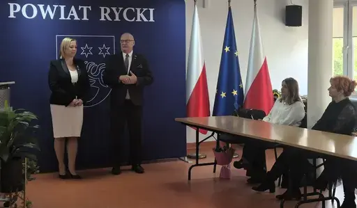 
                                                    Uroczystość w Starostwie Powiatowym w Rykach. Nagrodzeni nauczyciele i pracownicy oświaty z kwiatami i dyplomami pozują z władzami powiatu na tle granatowej ścianki z napisem Powiat Rycki.
                                                