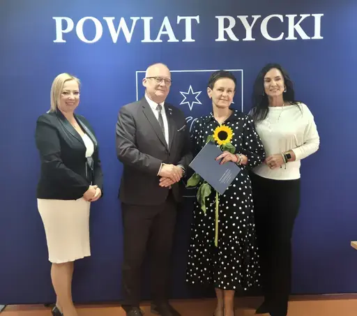 
                                                    Uroczystość w Starostwie Powiatowym w Rykach. Nagrodzeni nauczyciele i pracownicy oświaty z kwiatami i dyplomami pozują z władzami powiatu na tle granatowej ścianki z napisem Powiat Rycki.
                                                