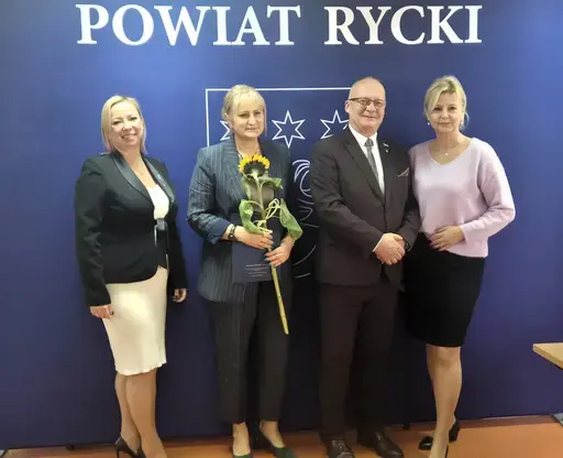 
                                                    Uroczystość w Starostwie Powiatowym w Rykach. Nagrodzeni nauczyciele i pracownicy oświaty z kwiatami i dyplomami pozują z władzami powiatu na tle granatowej ścianki z napisem Powiat Rycki.
                                                