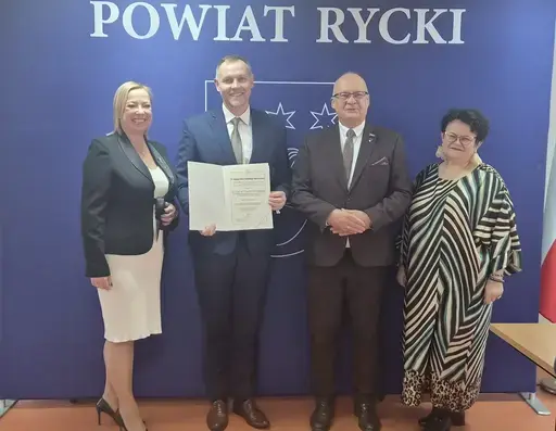 
                                                    Uroczystość w Starostwie Powiatowym w Rykach. Nagrodzeni nauczyciele i pracownicy oświaty z kwiatami i dyplomami pozują z władzami powiatu na tle granatowej ścianki z napisem Powiat Rycki.
                                                