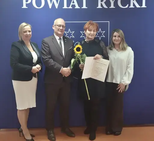 
                                                    Uroczystość w Starostwie Powiatowym w Rykach. Nagrodzeni nauczyciele i pracownicy oświaty z kwiatami i dyplomami pozują z władzami powiatu na tle granatowej ścianki z napisem Powiat Rycki.
                                                