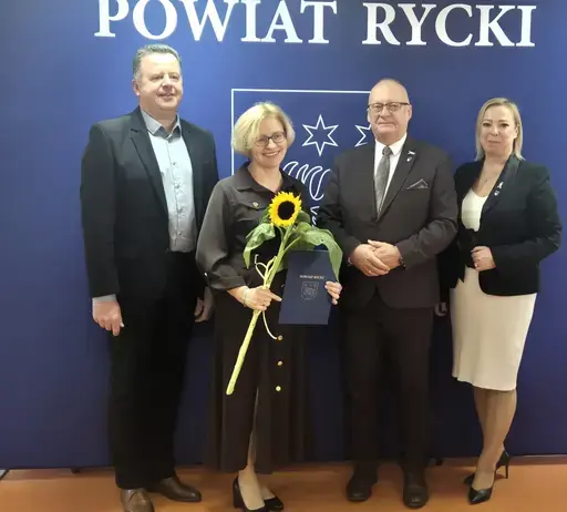 
                                                    Uroczystość w Starostwie Powiatowym w Rykach. Nagrodzeni nauczyciele i pracownicy oświaty z kwiatami i dyplomami pozują z władzami powiatu na tle granatowej ścianki z napisem Powiat Rycki.
                                                