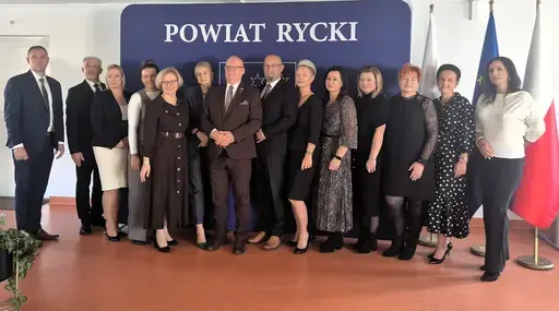 
                                                    Uroczystość w Starostwie Powiatowym w Rykach. Nagrodzeni nauczyciele i pracownicy oświaty z kwiatami i dyplomami pozują z władzami powiatu na tle granatowej ścianki z napisem Powiat Rycki.
                                                