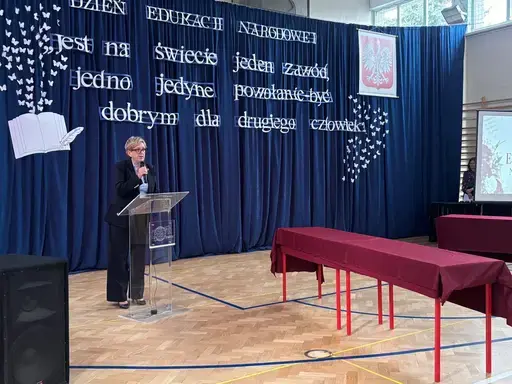 
                                                    Akademia z okazji Dnia Edukacji Narodowej w szkolnej sali gimnastycznej. Na granatowej kotarze napis: Jest na świecie jeden zawód, jedno jedyne powołanie – być dobrym dla drugiego człowieka. Uczniowie wręczają nauczycielom dyplomy i gratulacje.
                                                