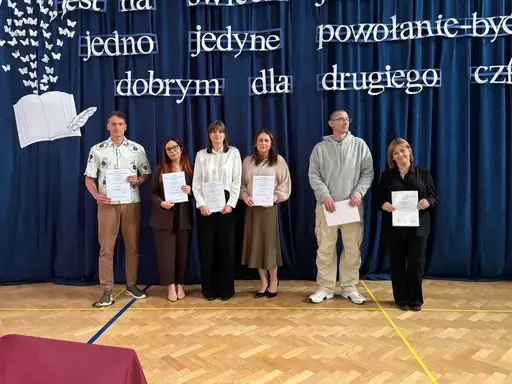 
                                                    Akademia z okazji Dnia Edukacji Narodowej w szkolnej sali gimnastycznej. Na granatowej kotarze napis: Jest na świecie jeden zawód, jedno jedyne powołanie – być dobrym dla drugiego człowieka. Uczniowie wręczają nauczycielom dyplomy i gratulacje.
                                                