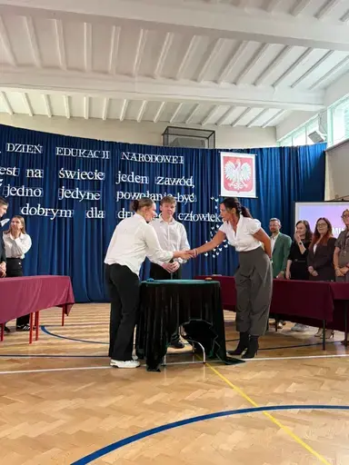 
                                                    Akademia z okazji Dnia Edukacji Narodowej w szkolnej sali gimnastycznej. Na granatowej kotarze napis: Jest na świecie jeden zawód, jedno jedyne powołanie – być dobrym dla drugiego człowieka. Uczniowie wręczają nauczycielom dyplomy i gratulacje.
                                                