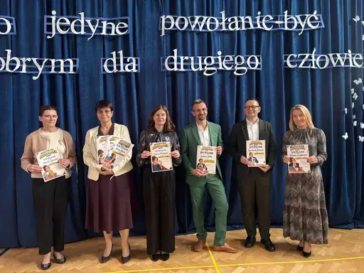 
                                                    Akademia z okazji Dnia Edukacji Narodowej w szkolnej sali gimnastycznej. Na granatowej kotarze napis: Jest na świecie jeden zawód, jedno jedyne powołanie – być dobrym dla drugiego człowieka. Uczniowie wręczają nauczycielom dyplomy i gratulacje.
                                                