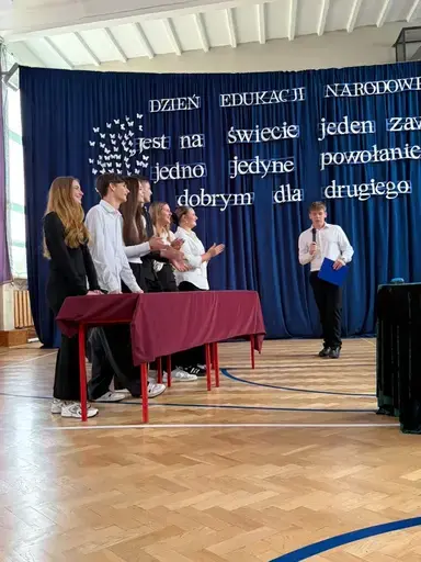 
                                                    Akademia z okazji Dnia Edukacji Narodowej w szkolnej sali gimnastycznej. Na granatowej kotarze napis: Jest na świecie jeden zawód, jedno jedyne powołanie – być dobrym dla drugiego człowieka. Uczniowie wręczają nauczycielom dyplomy i gratulacje.
                                                