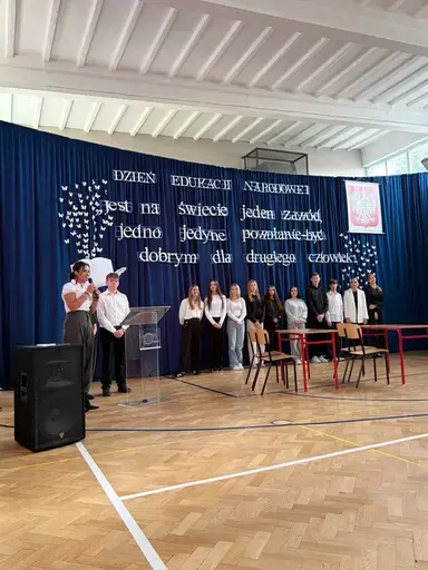
                                                    Akademia z okazji Dnia Edukacji Narodowej w szkolnej sali gimnastycznej. Na granatowej kotarze napis: Jest na świecie jeden zawód, jedno jedyne powołanie – być dobrym dla drugiego człowieka. Uczniowie wręczają nauczycielom dyplomy i gratulacje.
                                                