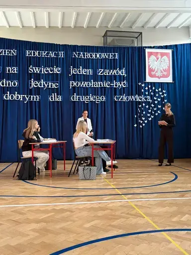 
                                                    Akademia z okazji Dnia Edukacji Narodowej w szkolnej sali gimnastycznej. Na granatowej kotarze napis: Jest na świecie jeden zawód, jedno jedyne powołanie – być dobrym dla drugiego człowieka. Uczniowie wręczają nauczycielom dyplomy i gratulacje.
                                                