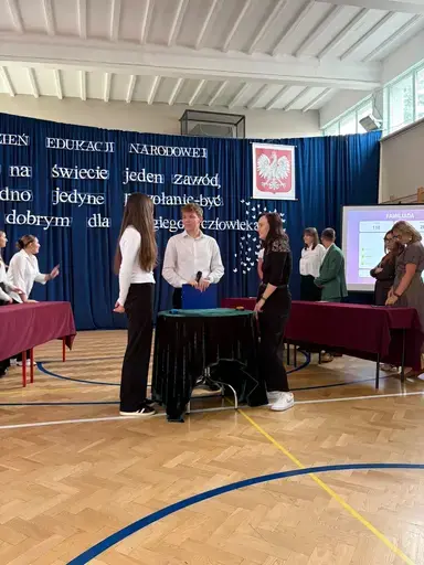 
                                                    Akademia z okazji Dnia Edukacji Narodowej w szkolnej sali gimnastycznej. Na granatowej kotarze napis: Jest na świecie jeden zawód, jedno jedyne powołanie – być dobrym dla drugiego człowieka. Uczniowie wręczają nauczycielom dyplomy i gratulacje.
                                                