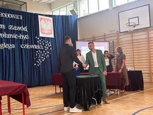 
                                                    Akademia z okazji Dnia Edukacji Narodowej w szkolnej sali gimnastycznej. Na granatowej kotarze napis: Jest na świecie jeden zawód, jedno jedyne powołanie – być dobrym dla drugiego człowieka. Uczniowie wręczają nauczycielom dyplomy i gratulacje.
                                                