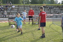 Chłopiec biegnący za piłką, grupa dzieci, trener, w tle ludzi na trybunach