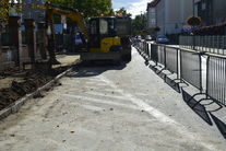 Zdjęcie przedstawia chodnik w trakcie remontu.Z prawej strony zabezpieczony pachołkami od ulicy. Na wprost widać koparkę, po lewej remontowany chodnik .