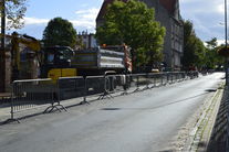 Zdjęcie przedstawia chodnik w trakcie remontu.Z lewej strony zabezpieczony pachołkami i barierkami od ulicy. Za nimi widać maszyny ciężkie,