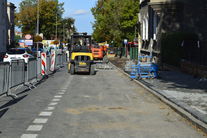 Zdjęcie przedstawia chodnik w trakcie remontu.Z lewej strony zabezpieczony pachołkami i barierkami od ulicy. Na wprost widać maszyny ciężkie, po prawej remontowany chodnik .