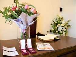 Leżące na biurku medale, kwiaty w wazonie oraz dyplomy z życzeniami.