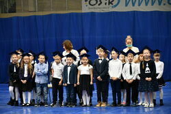 Dzieci z klasy pierwszej stojące w grupie w granatowych czapkach żaka na apelu w hali.