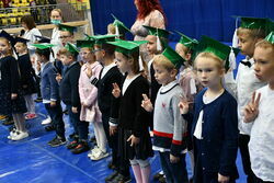 Dzieci z klasy pierwszej stojące w grupie w zielonych czapkach żaka na apelu w hali.