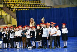 Dzieci z klasy pierwszej stojące w grupie w czerwonych czapkach żaka na apelu w hali.