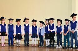 Grupa dzieci w akademickich czapkach ubranych na galowo stoi na tle jasnej ściany 