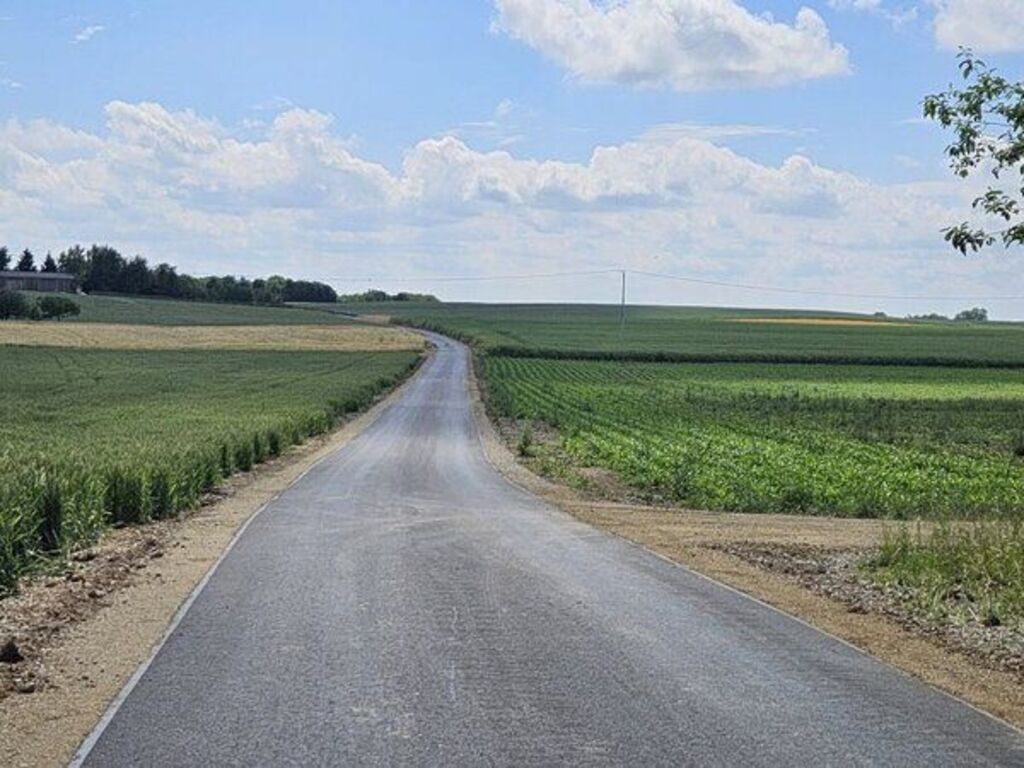 
                                                    Nowo wybudowana asfaltowa droga przecina pola uprawne i zielone tereny, w tle widoczne niebo z chmurami.
                                                