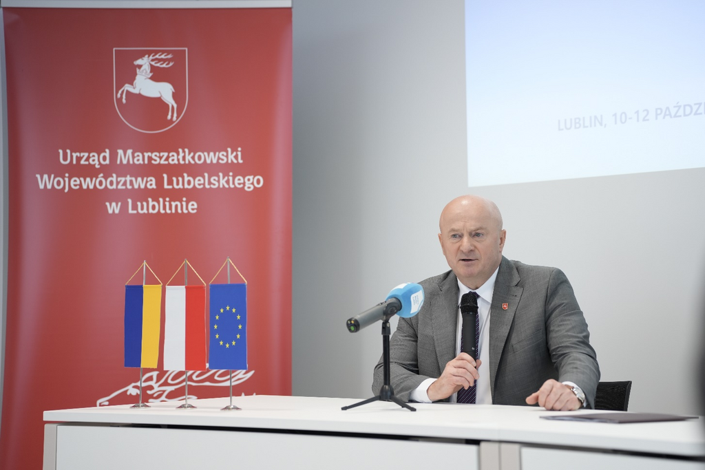 Mężczyzna w garniturze przemawia przy mikrofonie na konferencji, w tle banery z logo Urzędu Marszałkowskiego Województwa Lubelskiego i flagami Unii Europejskiej.