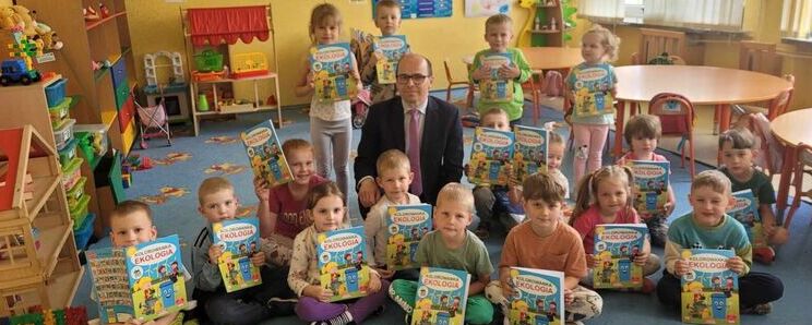 Grupa dzieci w przedszkolu razem z dorosłym mężczyzną. Dzieci trzymają książki, a pomieszczenie jest kolorowe i pełne zabawek.