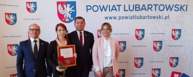 Czworo ludzi stoi na tle ścianki z napisem "Powiat Lubartowski" i herbem. Jedna kobieta trzyma dyplom i różę. Wszyscy są elegancko ubrani i uśmiechają się do zdjęcia.