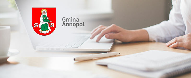 Osoba pracuje na laptopie z logo "Gmina Annopol" wyświetlonym na ekranie; biurko, notatnik i długopis obok.