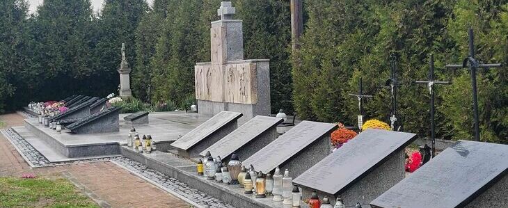 Opis zdjęcia: Pomnik i mogiły na cmentarzu wojskowym, z zniczami i kwiatami, otoczony drzewami.