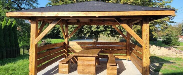 Drewniany altan ogrodowy z prostym, czarnym dachem, ławkami i stołem, umiejscowiony na trawniku w słoneczny dzień.