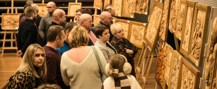 Grupa osób ogląda obrazy na wystawie sztuki w galerii z drewnianymi ramami ustawionymi w rzędach.