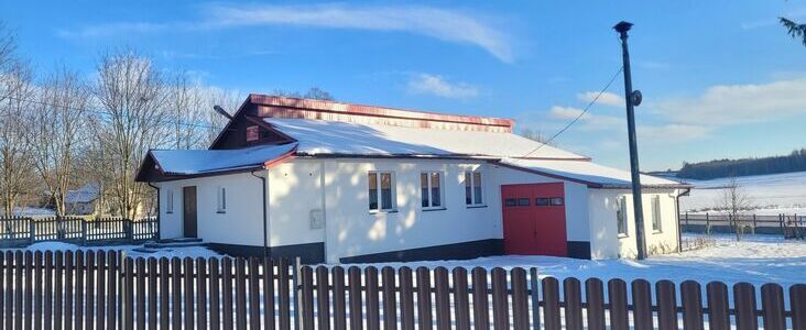 Mały biały dom z czerwonymi okiennicami i dachem, stojący przed zaśnieżonym polem, oddzielony brązowym drewnianym płotem, z kominkiem wydobywającym dym na tle bezchmurnego nieba.