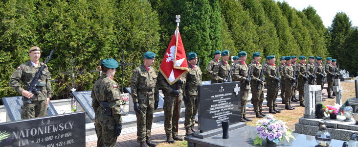 Grupa żołnierzy w mundurach stoi na cmentarzu wojskowym wzdłuż rzędu nagrobków, z flagą kraju wśród nich. To uroczystość lub ceremonia.