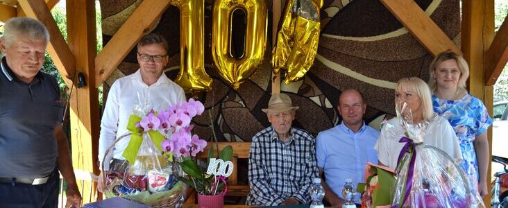 Pięć osób pozujących ze starszym mężczyzną siedzącym przy stole z prezentami i balonami "100" świętującymi stulecie.